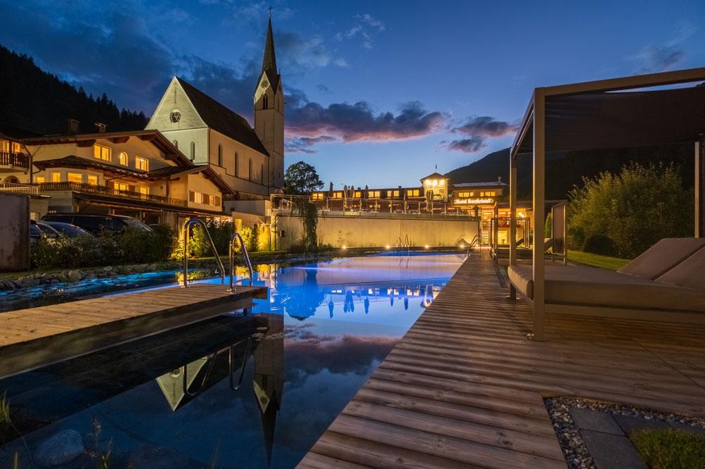 Hotelzwembad met houten terras bij schemering naast een kerk
