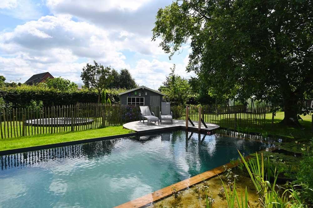 Natuurlijk zwembad met houten terras, ligstoelen en tuinhuis in groene tuin